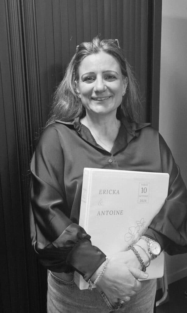 Livre de mariage Ericka & Antoine 10 octobre 2026 Une femme souriante en chemise noire et jean tient fermement un livre de mariage clair avec "ERICKA & ANTOINE" et "SAMEDI 10 OCTOBRE 2026".