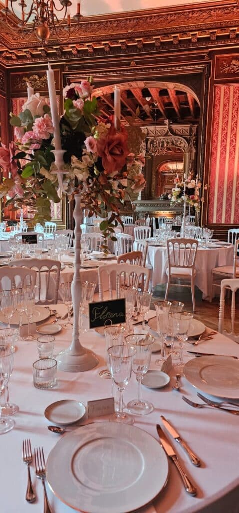 Grande table de réception élégante dressée avec vaisselle raffinée, fleurs dans un haut candélabre blanc, et chevalet 'Florence'. Décor intérieur somptueux.
