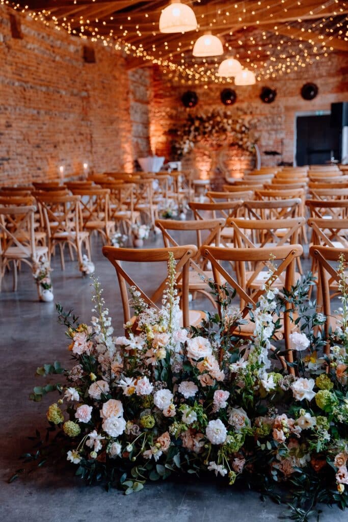 Décoration mariage rustique chic : ambiance florale et chaises bois. Décor de cérémonie de mariage dans une salle en briques : rangées de chaises en bois clair et grand arrangement floral, éclairage féerique.
