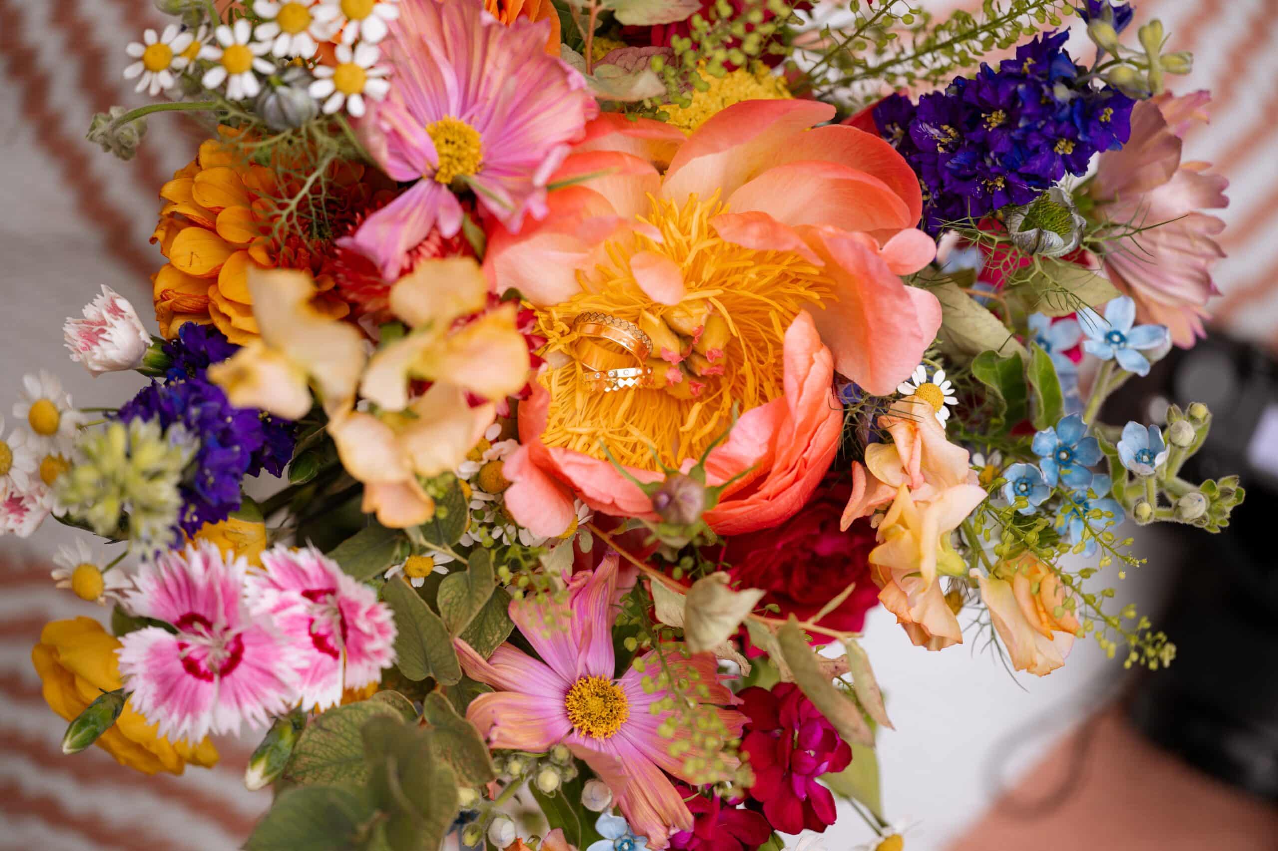 Deux alliances dorées nichées au cœur d'une grande pivoine corail et jaune, entourée d'un bouquet vibrant de fleurs variées.