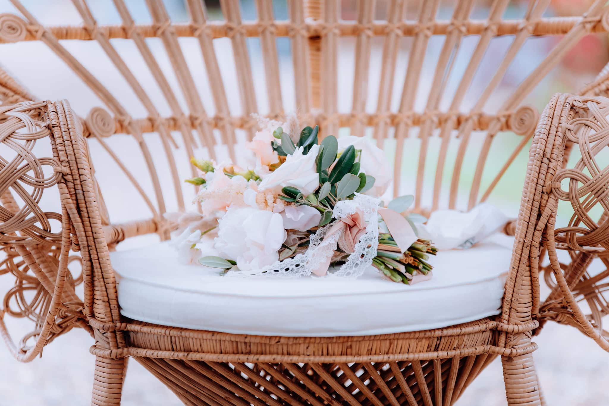 Bouquet nuptial pastel de pivoines, roses et feuillage, orné de dentelle et rubans, posé sur le coussin blanc d'une chaise en rotin.