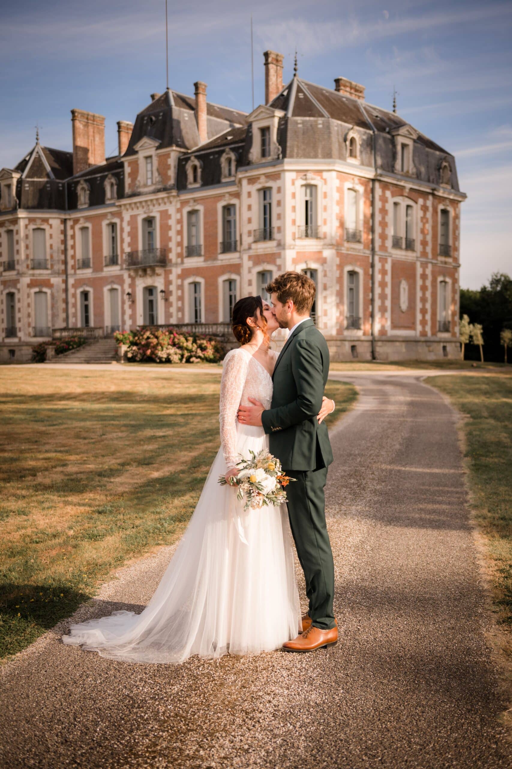 Mariés s'embrassant devant un château historique français en briques. La mariée porte une robe en dentelle; le marié un costume vert.