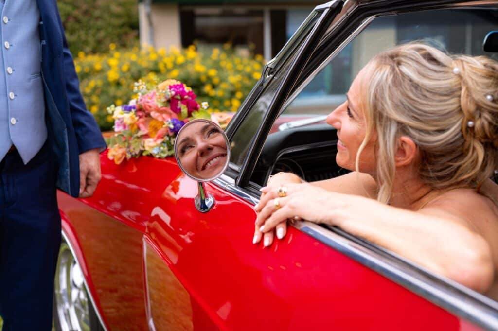 Mariage en décapotable rouge : le sourire éclatant de la mariée Mariée joyeuse dans une décapotable rouge, son sourire reflété dans le rétroviseur. Le marié en costume bleu et un bouquet sont visibles.